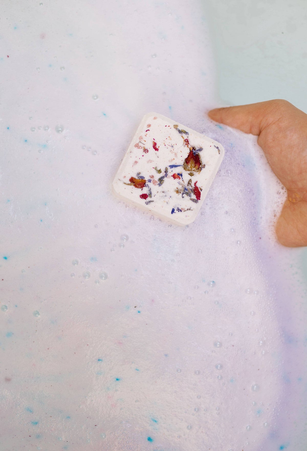 Square bath bomb cube floating in light purple water, with red and blue flower-like speckles