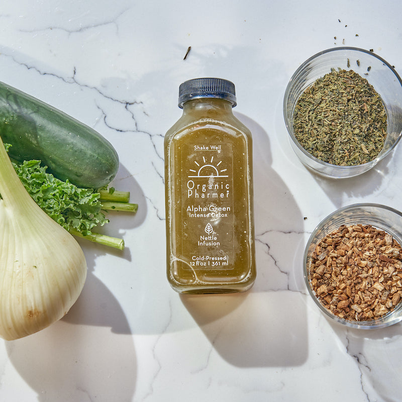 Green juice bottle with black cap, with herbs, seeds, and vegetables nearby