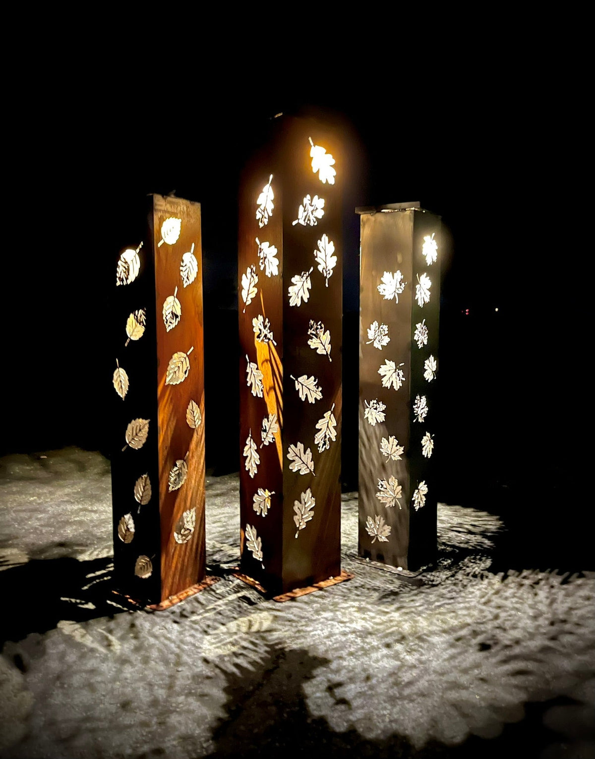 Set of lit solar pillar luminaries with etched leaf patterns on metal pillars
