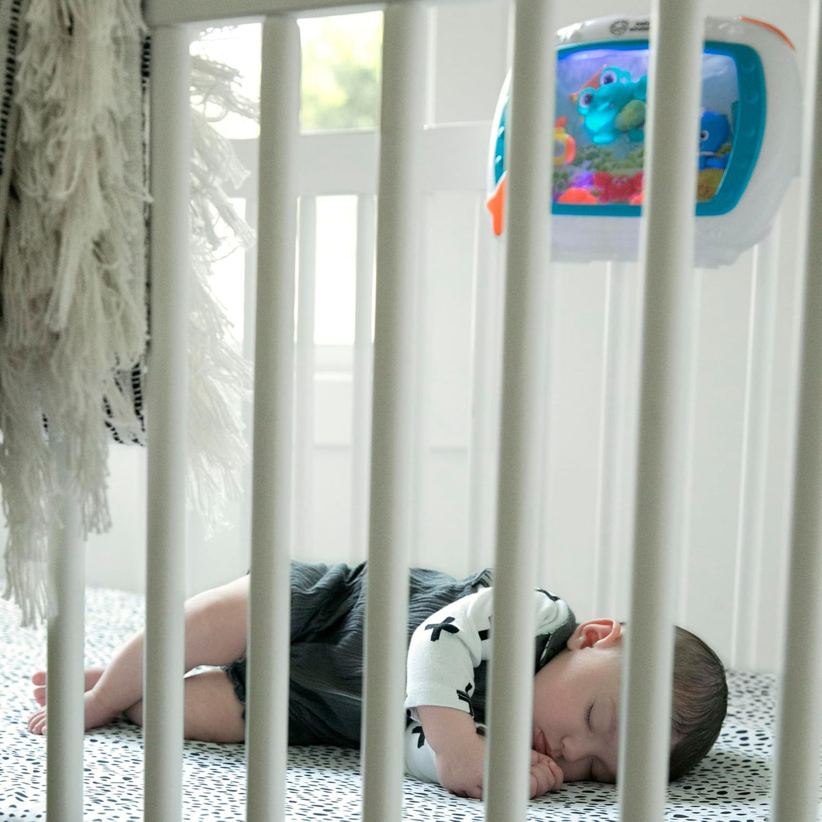 Baby lying next to blue crib toy and sound machine on patterned blanket