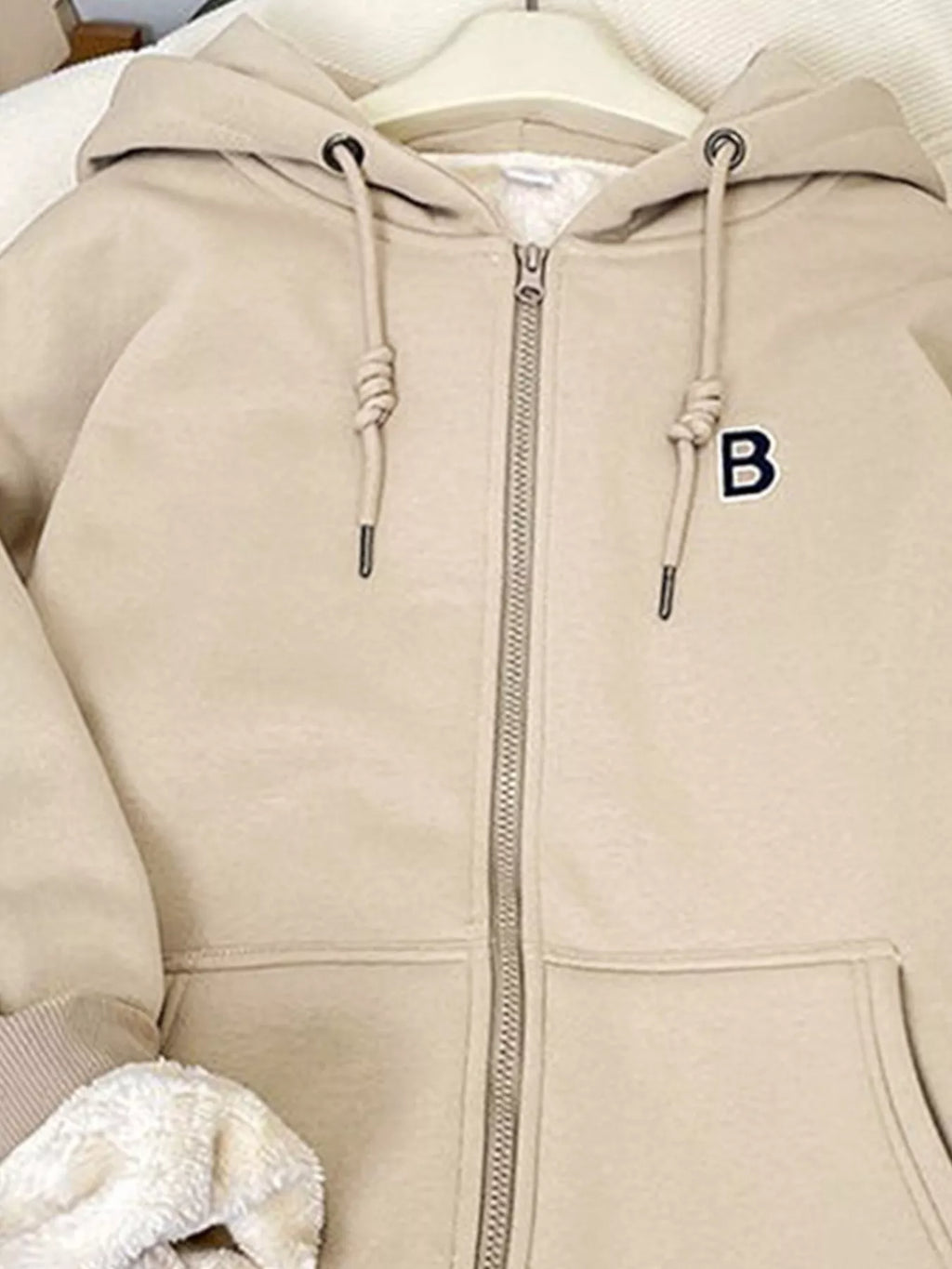Light beige fleece-lined zip-up hoodie close-up: hood, zipper, drawstring cords, pocket.