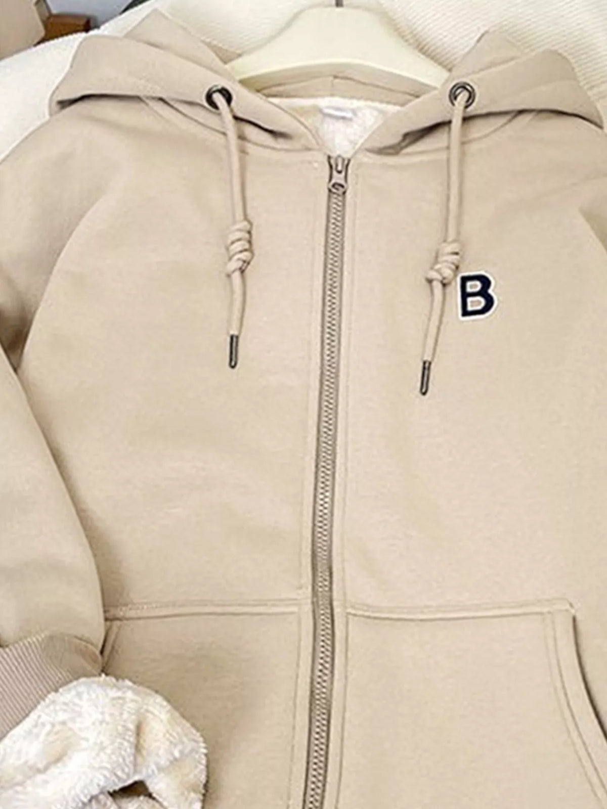 Light beige fleece-lined zip-up hoodie close-up: hood, zipper, drawstring cords, pocket.
