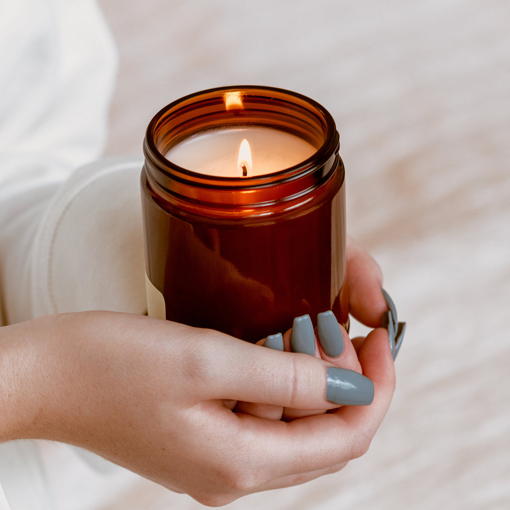 Amber glass candle jar with lit wick held in hands, brown wax and printed label