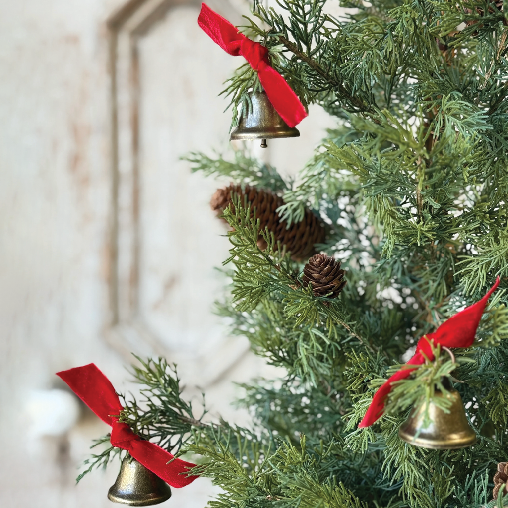 Decorative brass-tone bell ornament with red ribbon hanging from a pine tree.