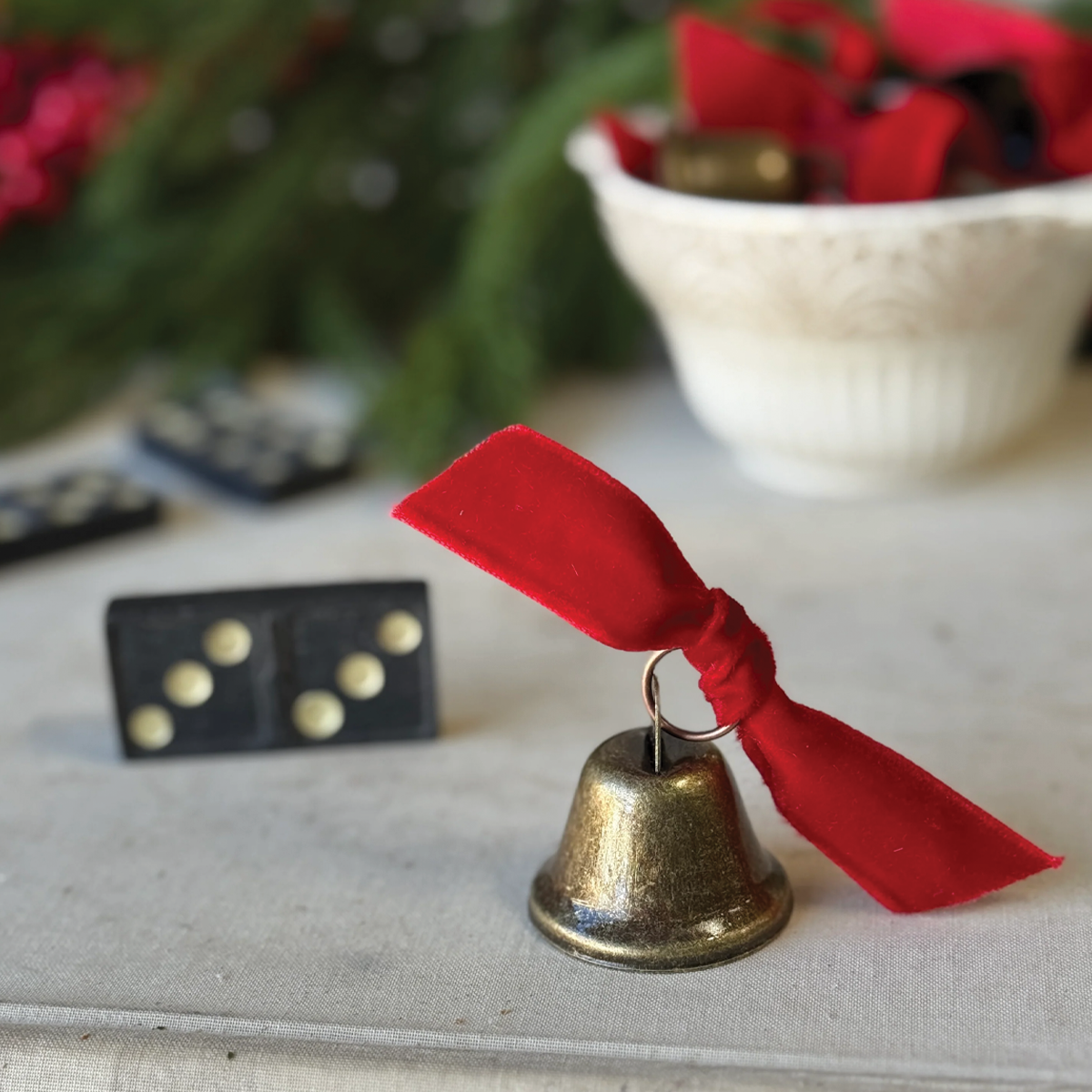 Decorative brass-tone bell ornament with red ribbon on white fabric near blurred greenery.