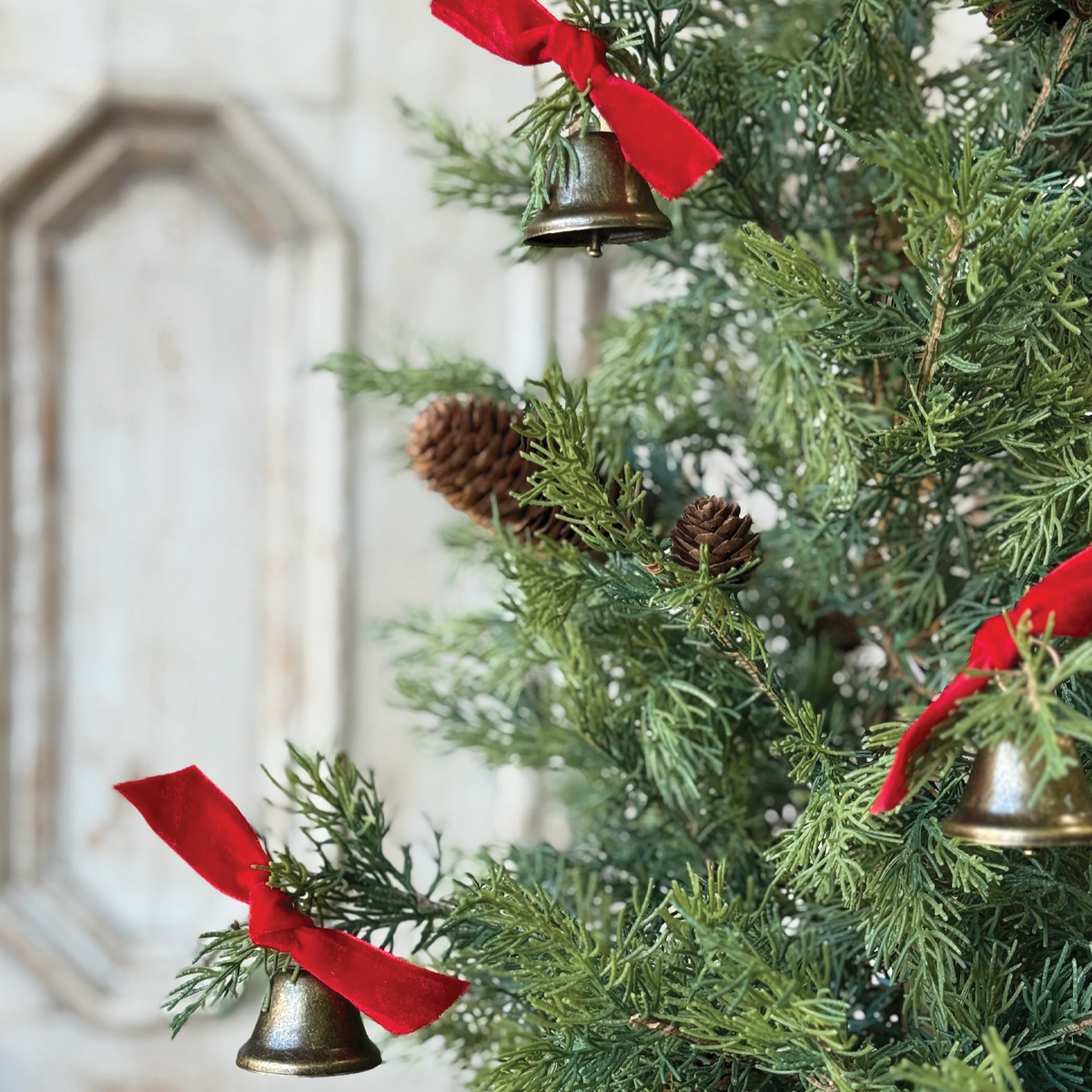 Decorative brass-tone bell ornament with red ribbon hanging from a pine tree.