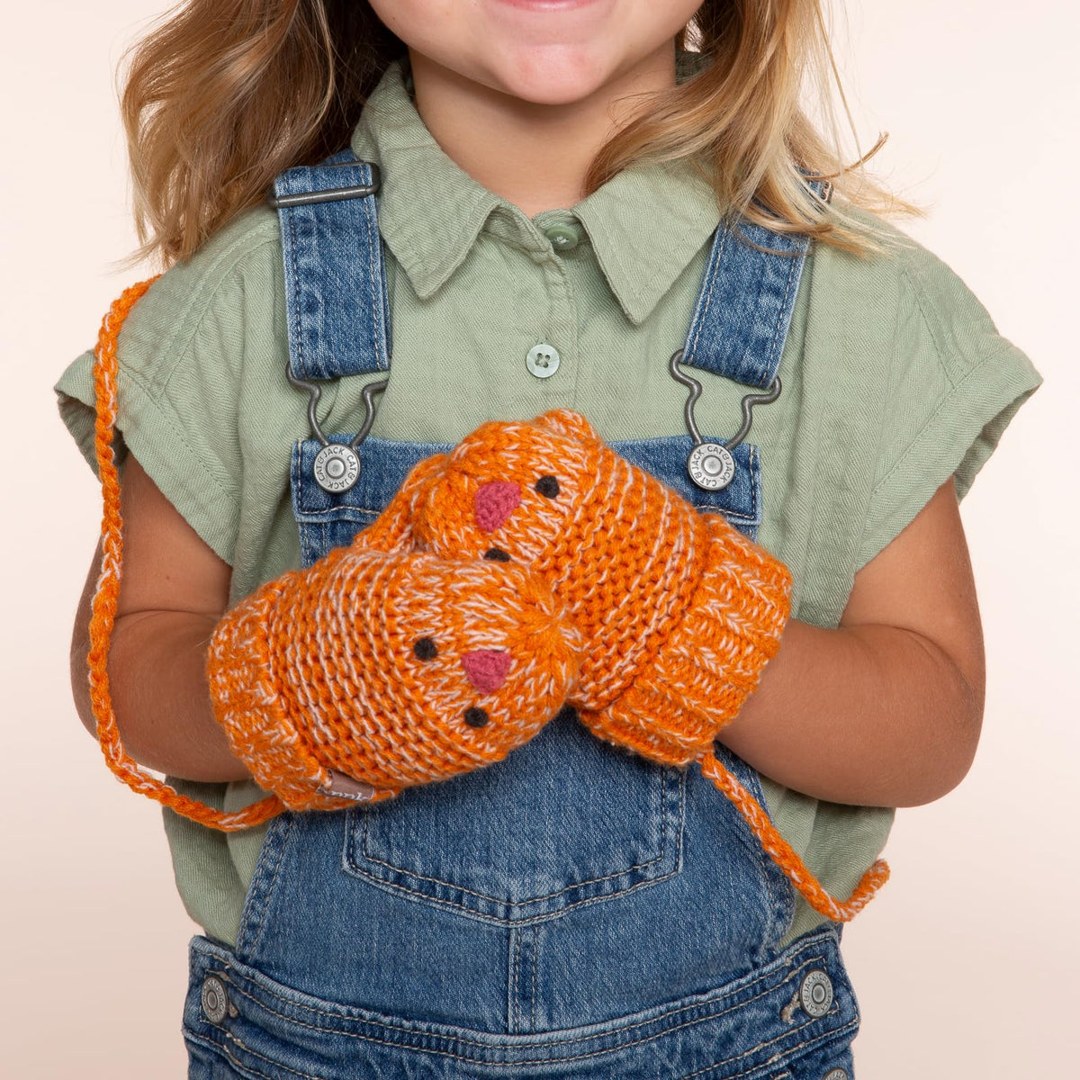Rusty orange chick mittens with cord, worn by a child on denim overalls