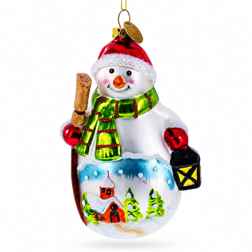 Glass Christmas ornament of snowman holding broom and lantern, red hat, green scarf