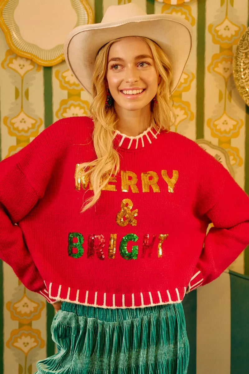 Red knit sweater with gold sequin “MERRY & BRIGHT” lettering and white fringe neckline