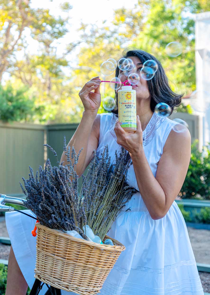 Woman holding chamomile & herbs bubble bath bottle while blowing bubbles outdoors.