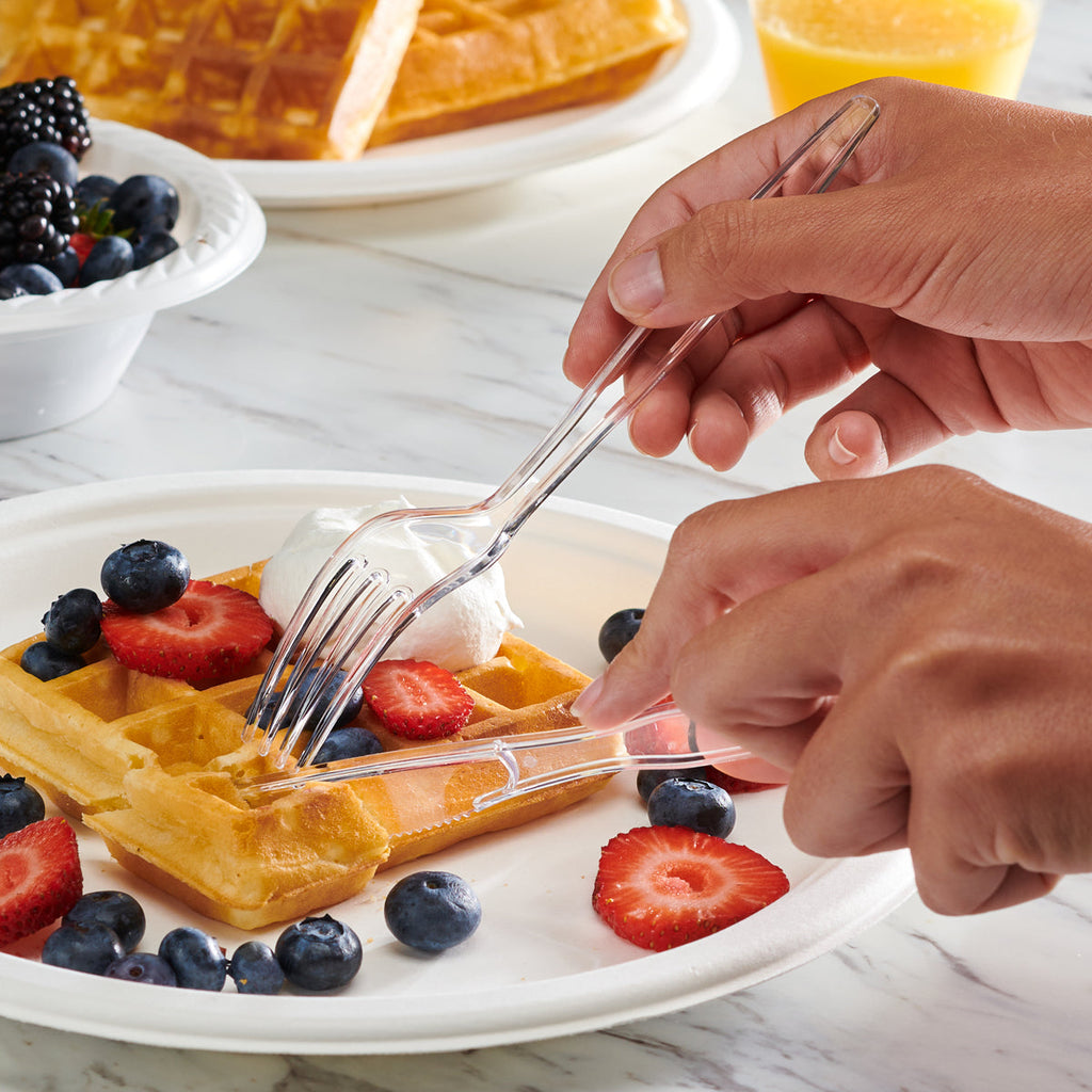 Hands using a clear plastic fork on a waffle topped with berries