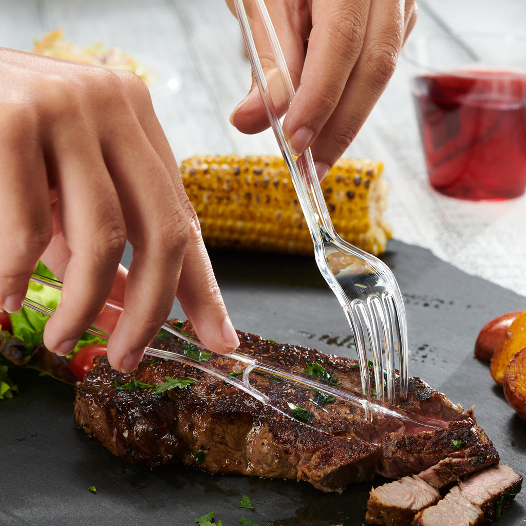Clear plastic fork in hands serving steak on a flat surface
