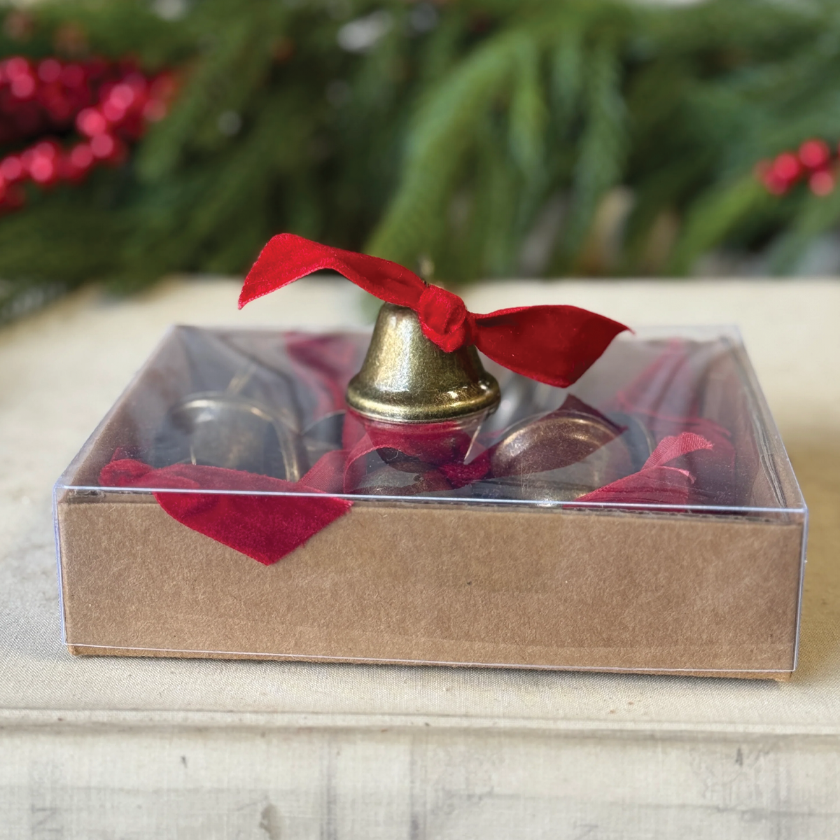 Clear gift box containing a decorative brass-tone bell with red ribbon bow.