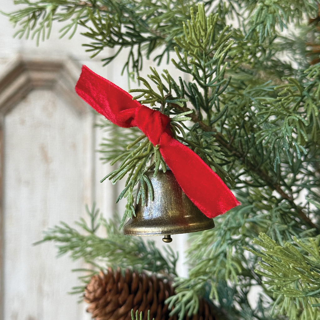 Decorative brass-tone bell ornament with red ribbon tied to a pine tree branch.