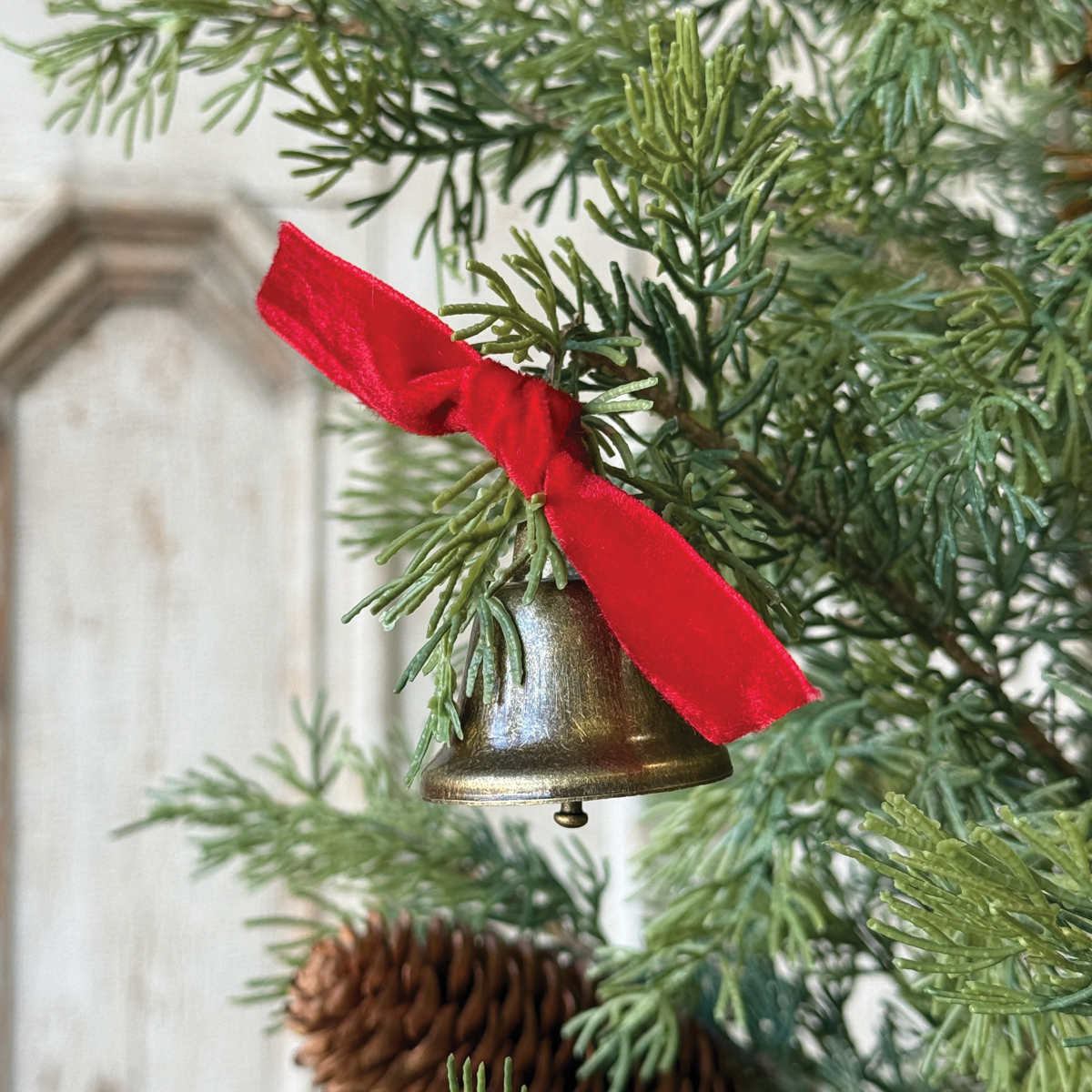 Decorative brass-tone bell ornament with red ribbon tied to a pine tree branch.