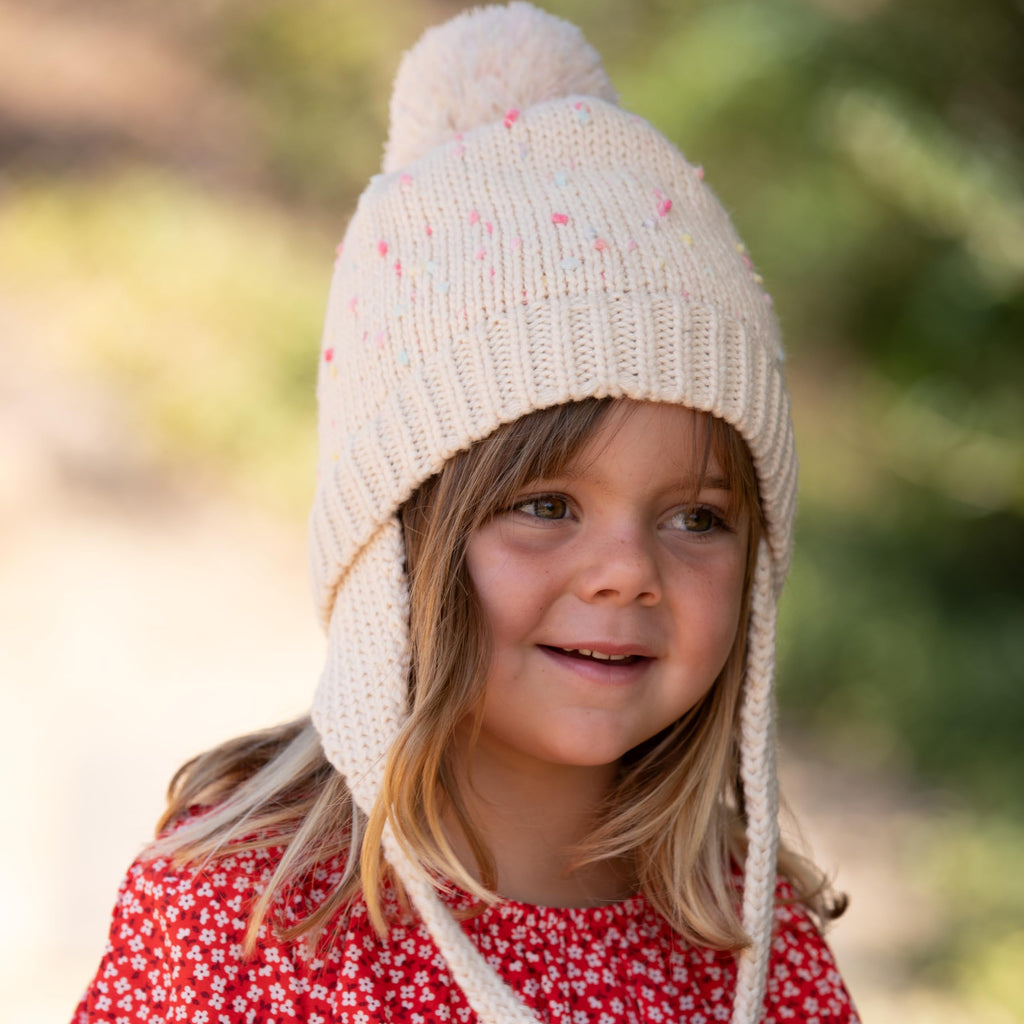 Off-white speckled knit earflap beanie with pom-pom and tie strings on a child.