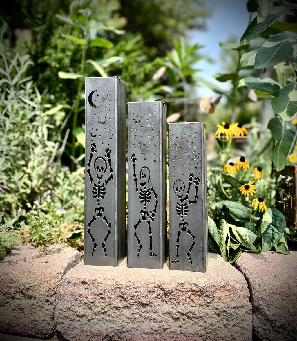 Three steel solar tube lights with skeleton silhouettes on each front panel in a garden