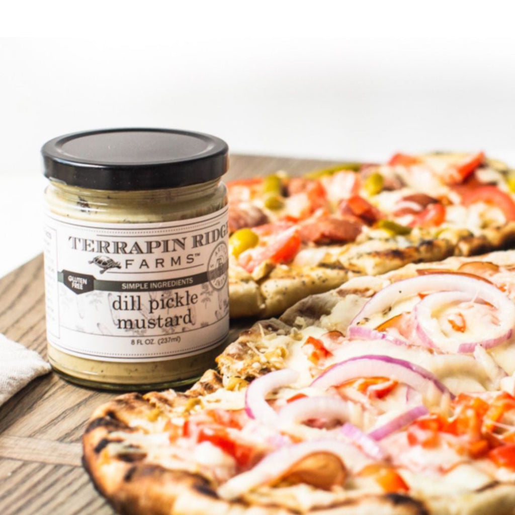 Jar of dill pickle mustard with black lid, shown with pizza slices