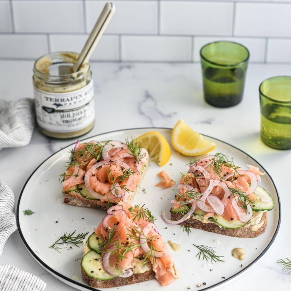 Jar of dill pickle mustard with black lid beside open-faced shrimp sandwich