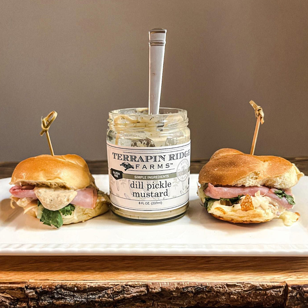 Jar of dill pickle mustard with black lid, flanked by two burgers