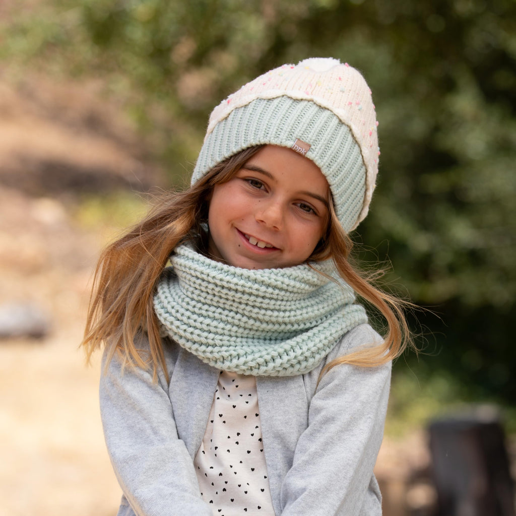 Toddler girl wearing a mint knit beanie and cream speckled scarf set outdoors.