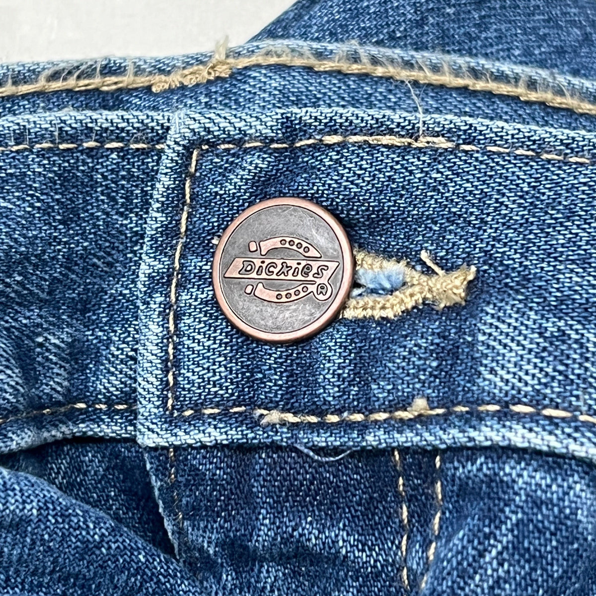 Riveted leather-look logo button on blue denim waistband of carpenter jeans.