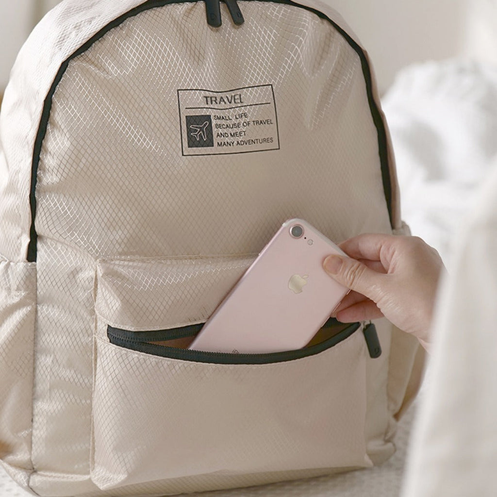 Beige foldable backpack front pocket with travel label and visible smartphone.