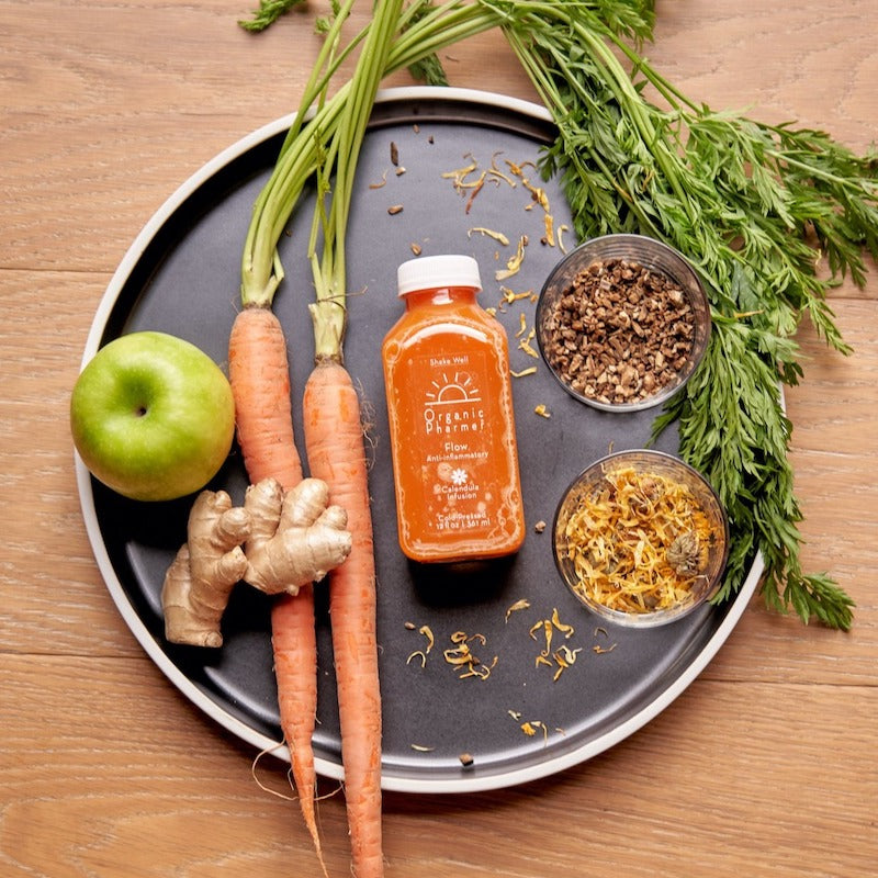 Organic beverage bottle on tray with carrots, ginger, apple, herbs, and dried botanicals.