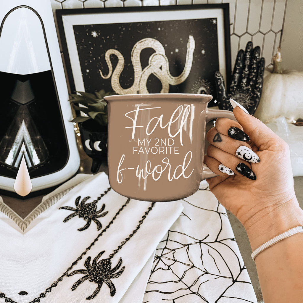 Brown ceramic coffee mug with white “fall” and “f-word” lettering held near decor