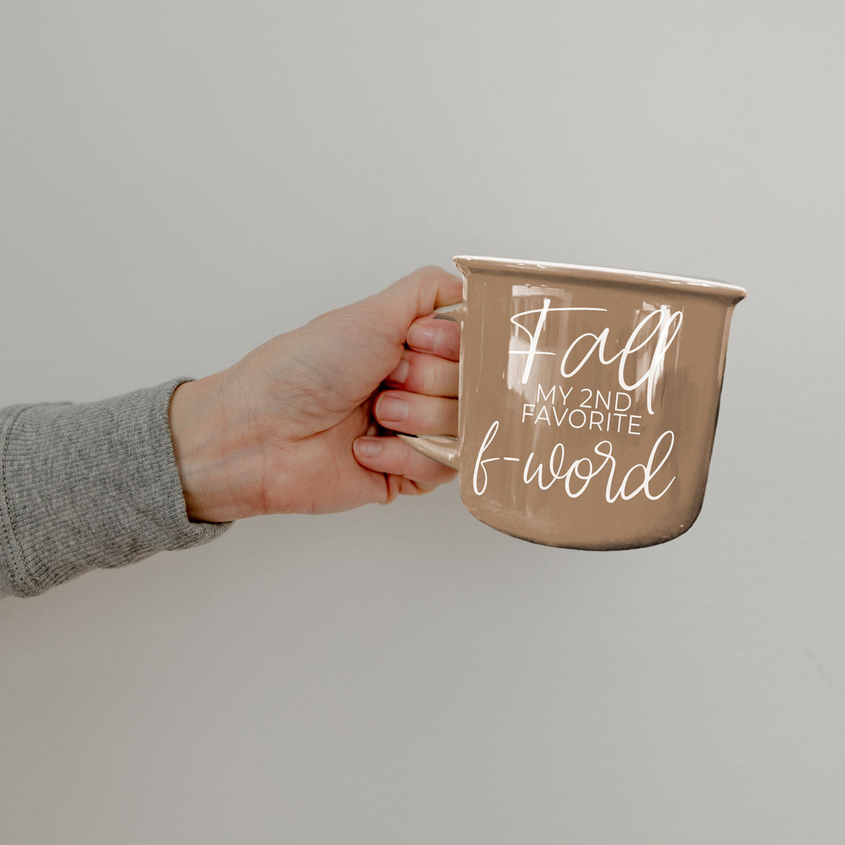 Brown ceramic coffee mug held in hand with white “fall” and “f-word” lettering