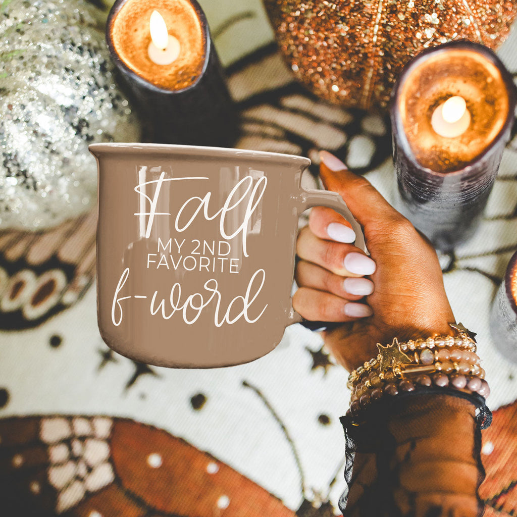 Brown ceramic mug with white “fall” and “f-word” lettering held outdoors with candles