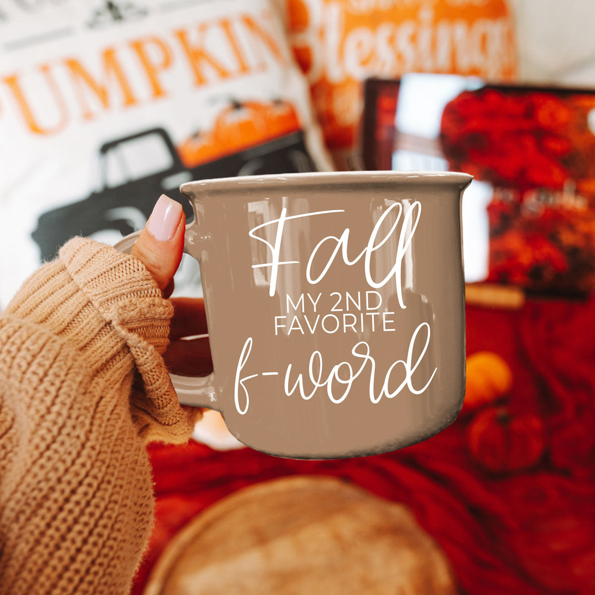 Beige mug with white lettering held in a person’s hand in front of autumn decor