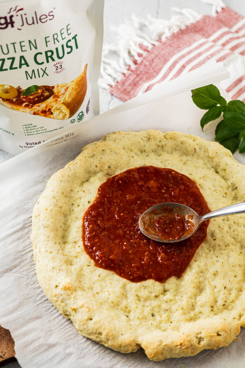 Prepared gluten-free pizza crust with red sauce center and a spoon spreading it
