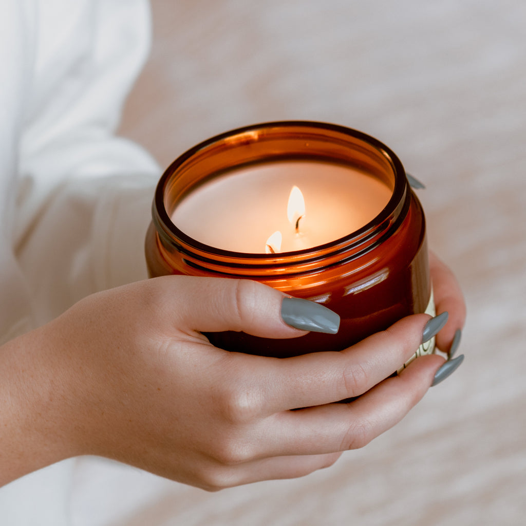 Amber glass candle jar with flame, held in hands with gray-blue nails