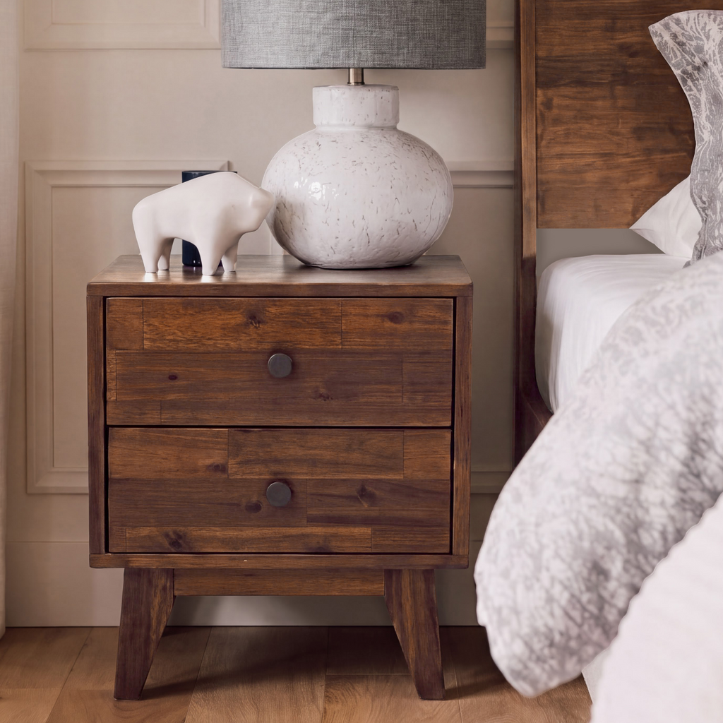 Solid wood nightstand with two drawers and round knobs next to a bed.