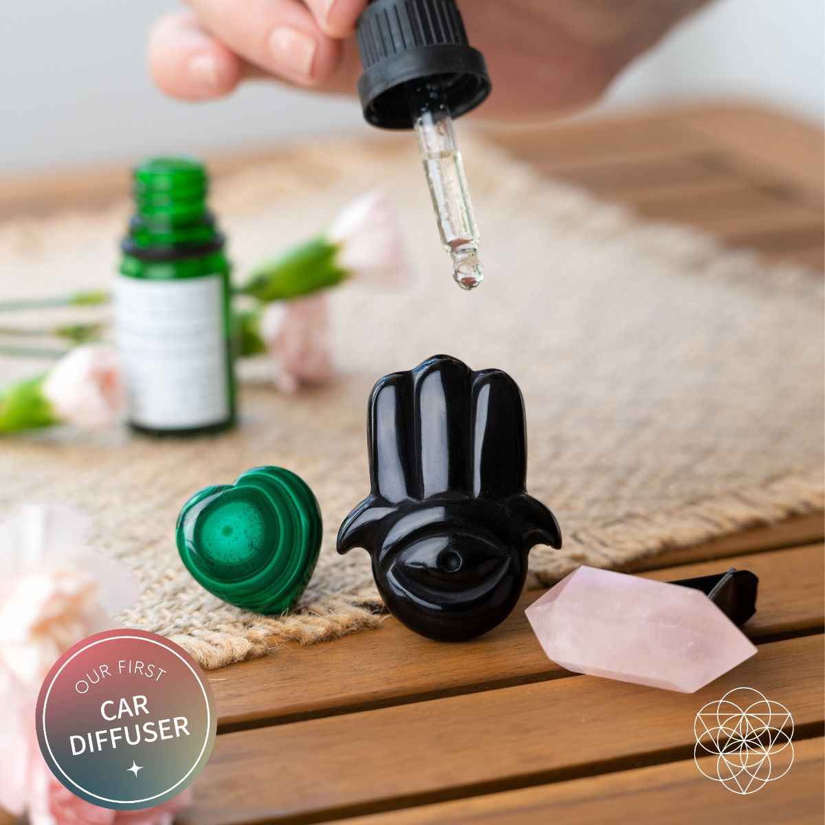 Aromatherapy dropper dispensing oil onto black hamsa car diffuser with pink and green crystals