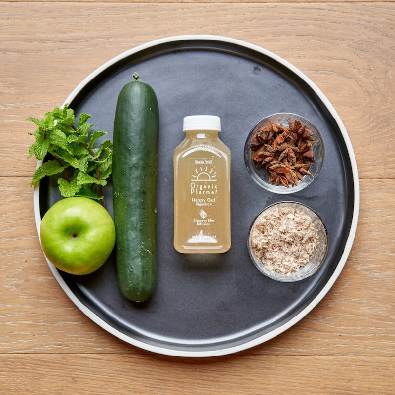 Black tray with small amber bottle, cucumber, apple, herbs, and dried brown flakes