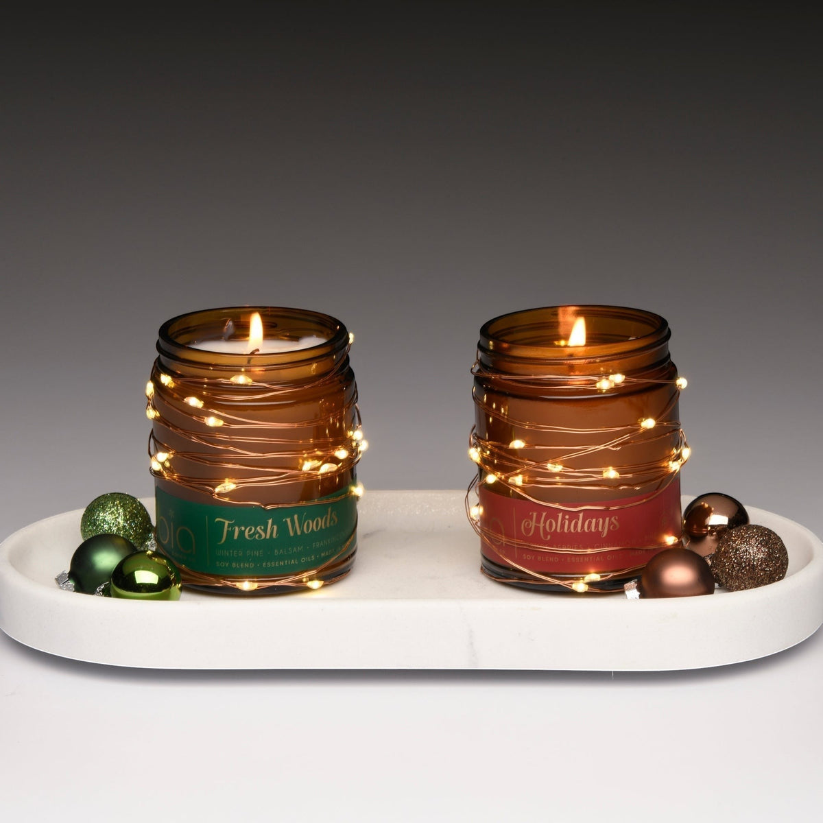 Two amber candle jars with lit wicks on a white tray; green and brown accents