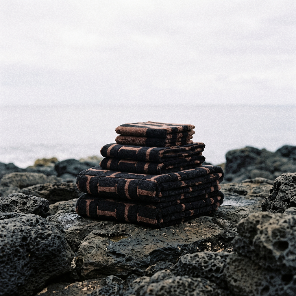 Folded cotton towels in tobacco and black stacked on dark rocks at the shoreline