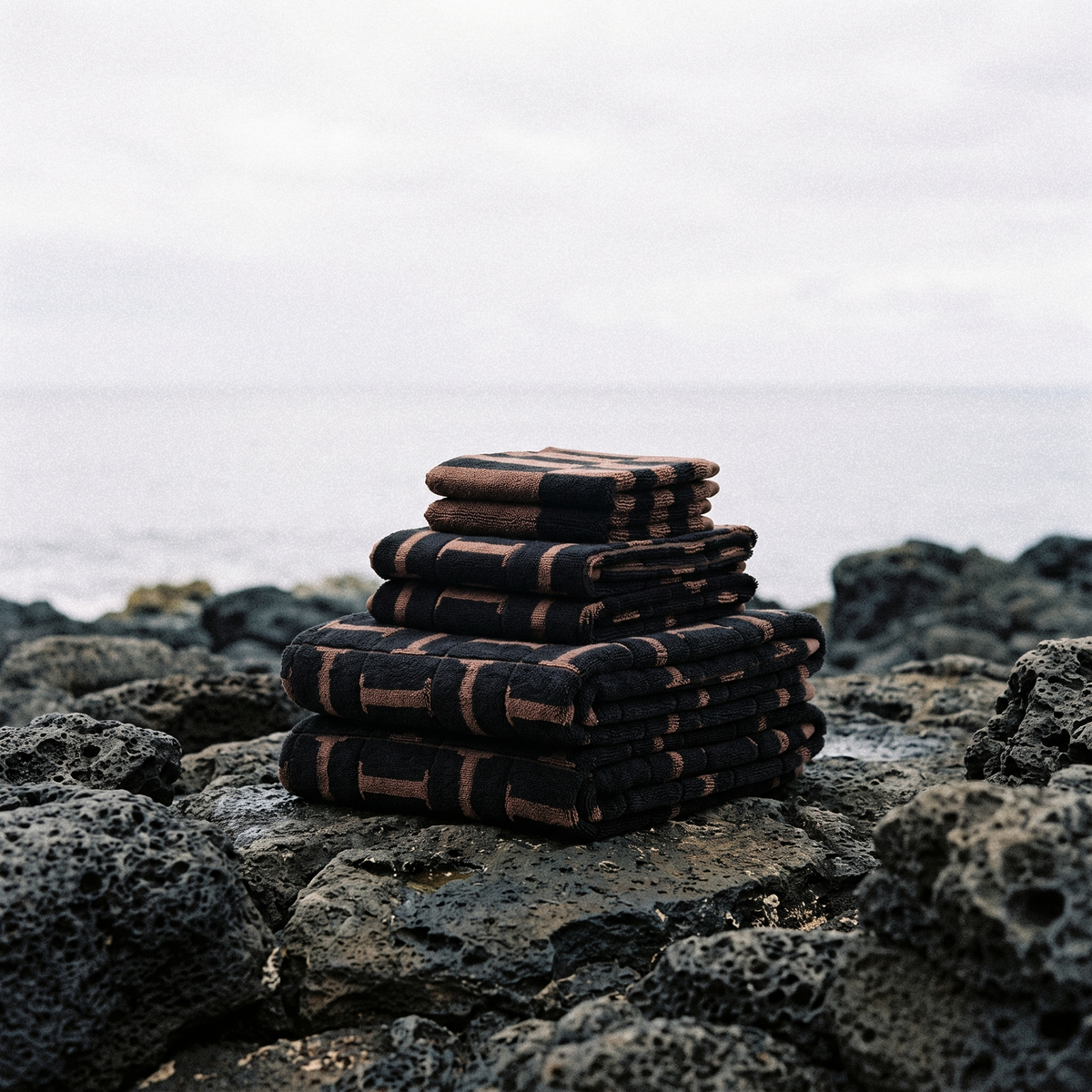 Folded cotton towels in tobacco and black stacked on dark rocks at the shoreline