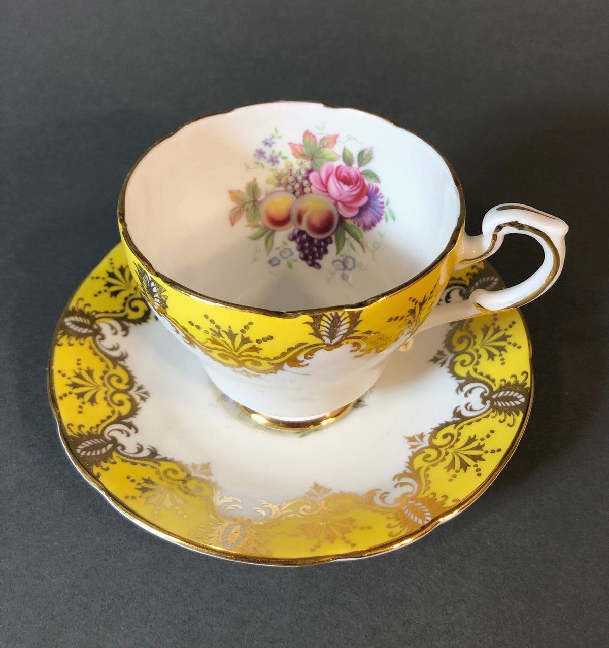 Yellow and gold porcelain tea cup with pink rose, grapes, and fruit bouquet on saucer