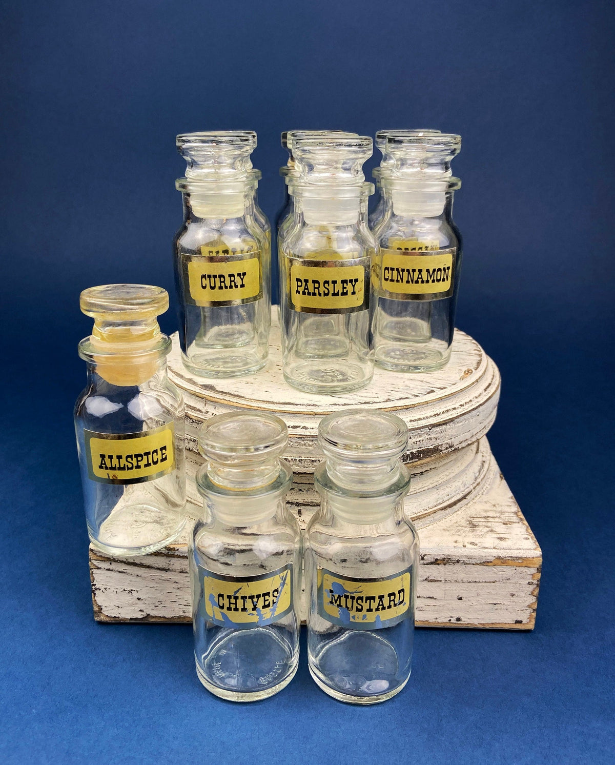 Small clear spice jars with gold text labels on stacked white wood blocks