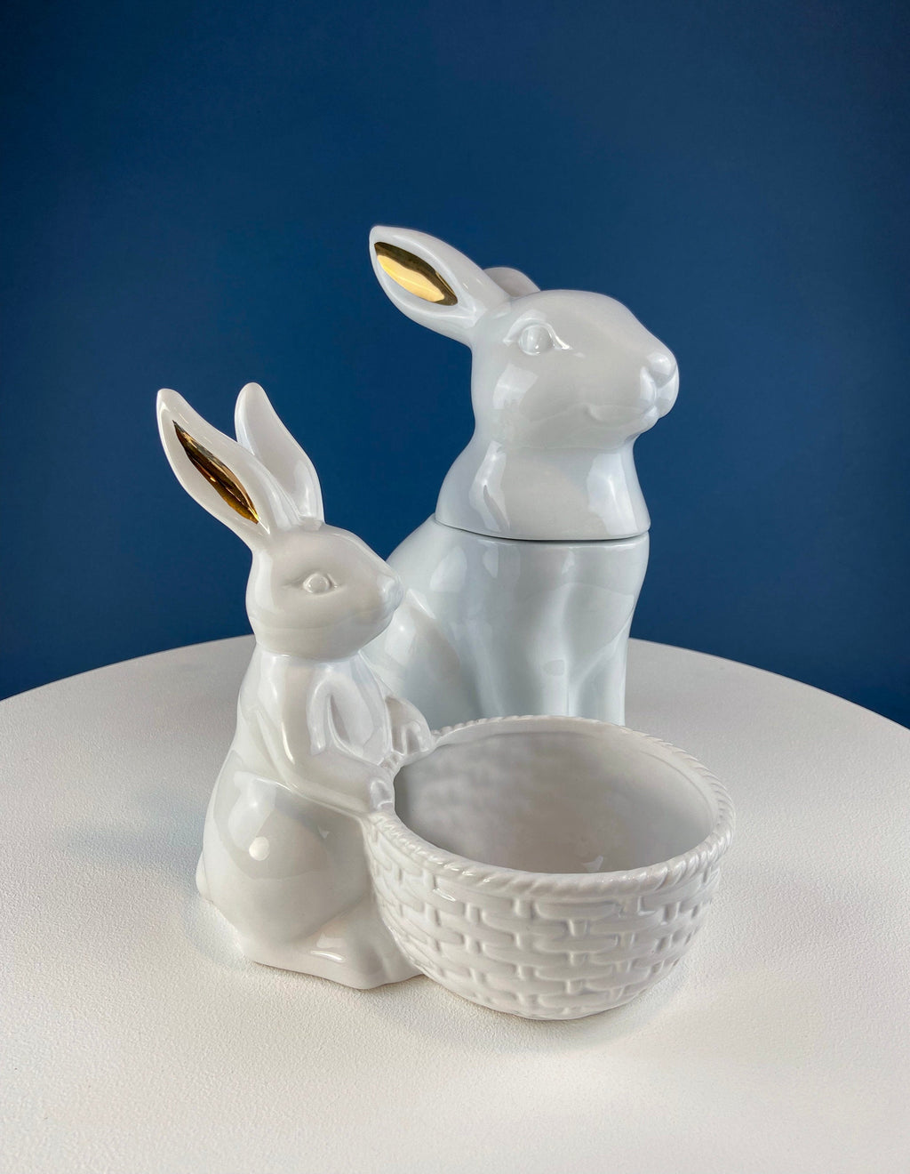 White ceramic rabbits beside a woven basket holding pastel speckled eggs