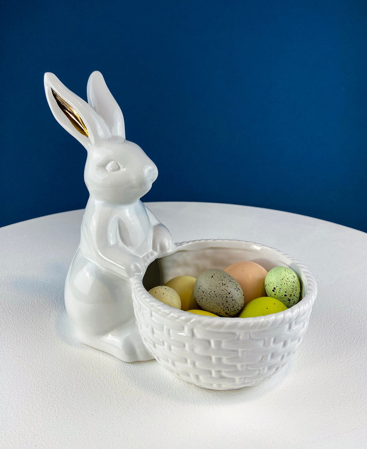 White ceramic rabbit holding an egg basket filled with pastel-colored eggs