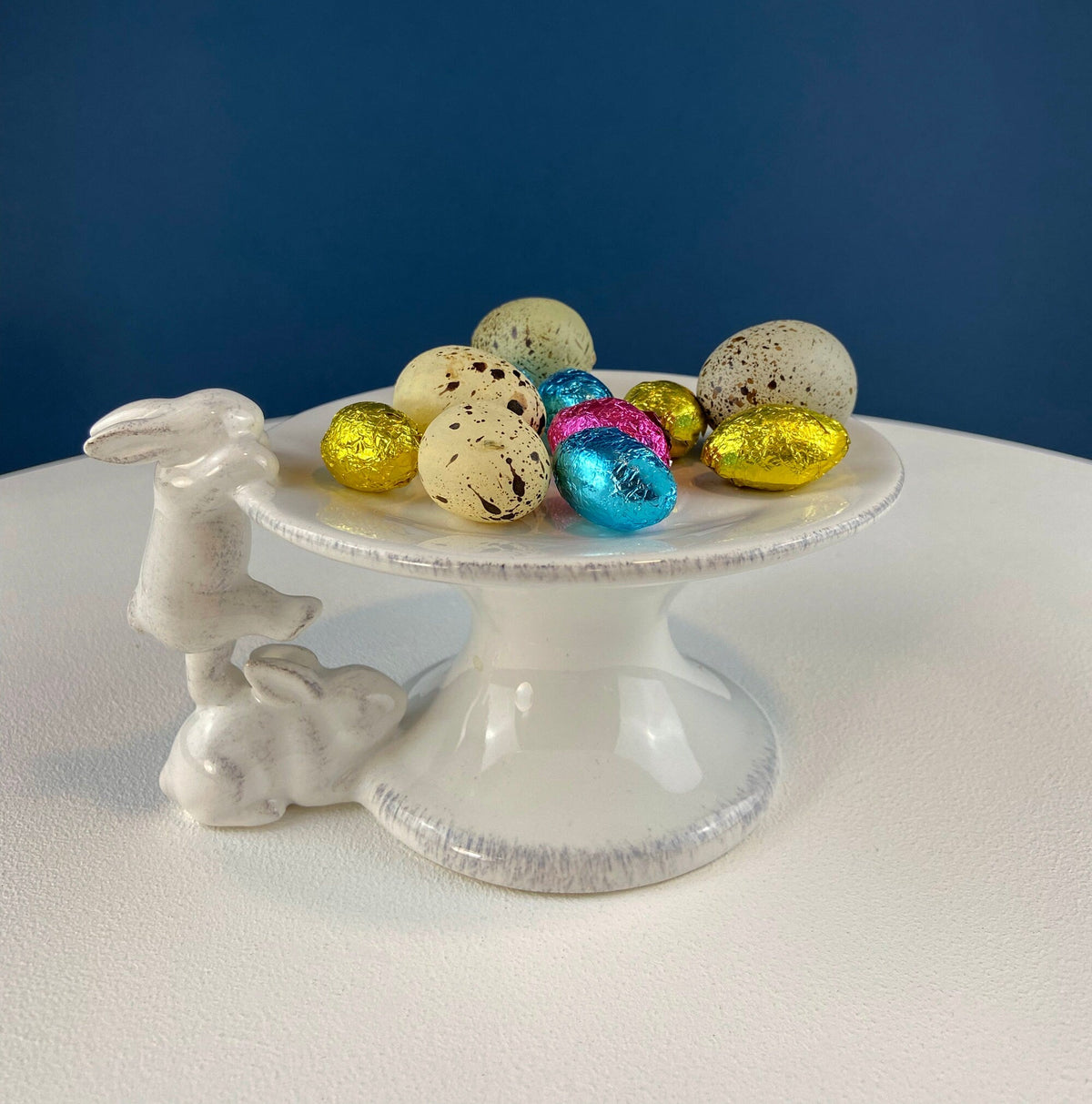 White ceramic bunny cupcake stand with round plate filled with colorful speckled eggs