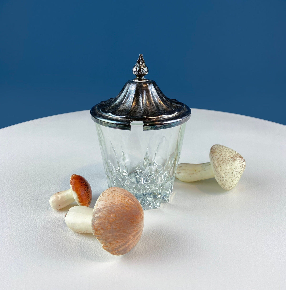 Square clear glass jar with ornate silver lid, with small serving utensils nearby