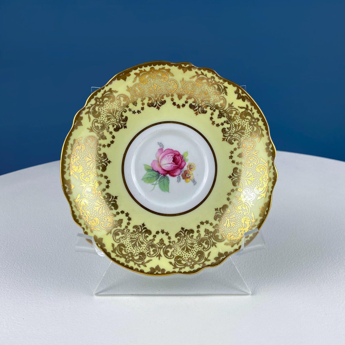 Yellow saucer with gold ornamental rim and pink rose center, displayed on clear stand