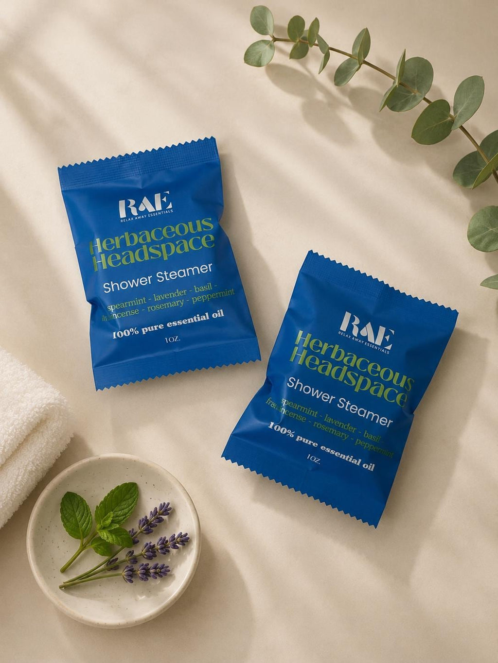 Two blue BAE shower steamer packets with herbal scent list, beside towel and dried herbs