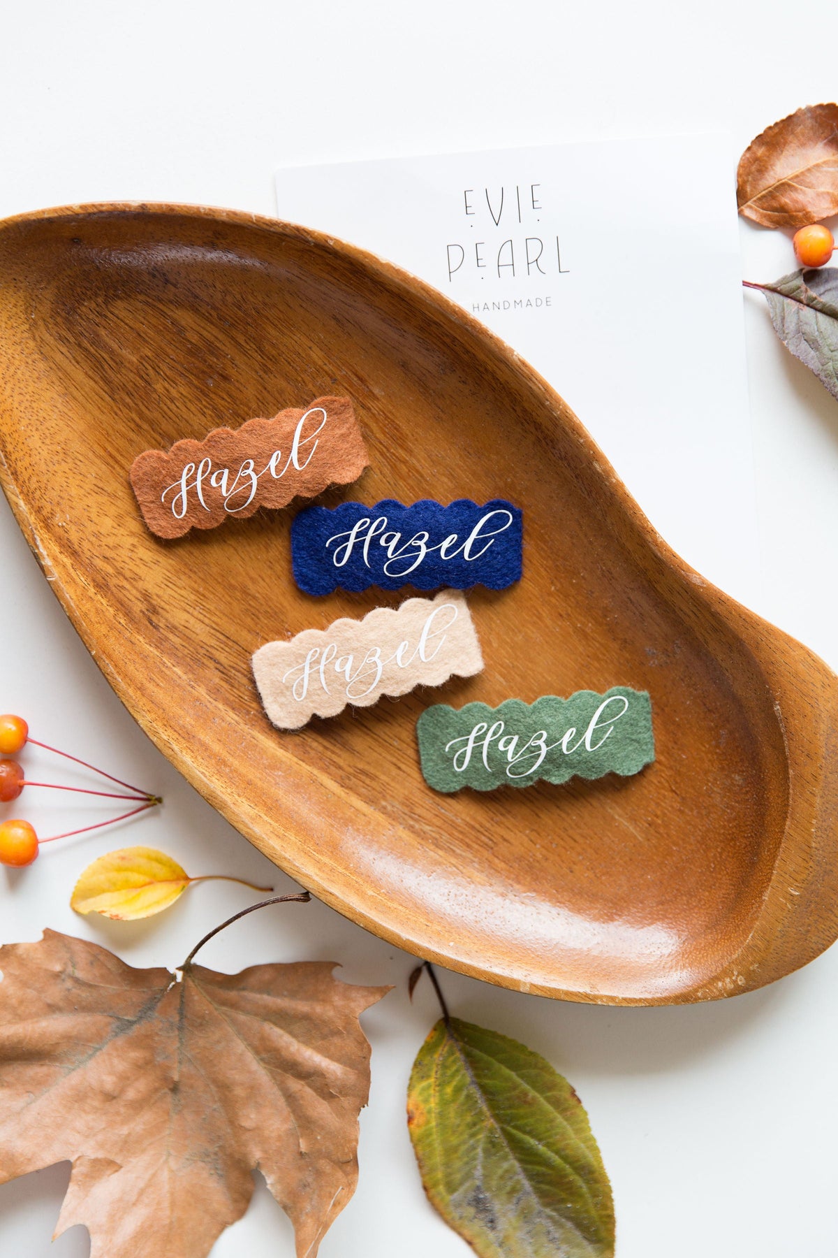 Felt snap hair clips with stitched names in blue, tan, and green on a wooden tray.