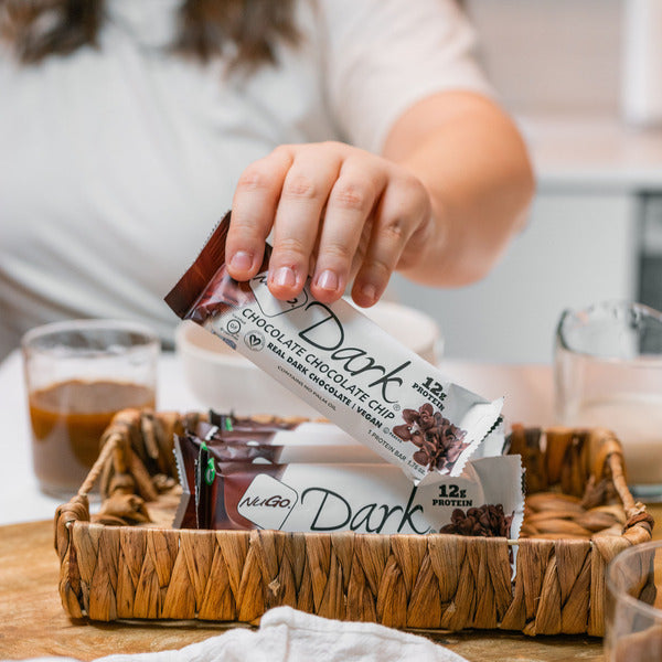 Person holding NuGo Dark chocolate chip protein bar over a basket of bars